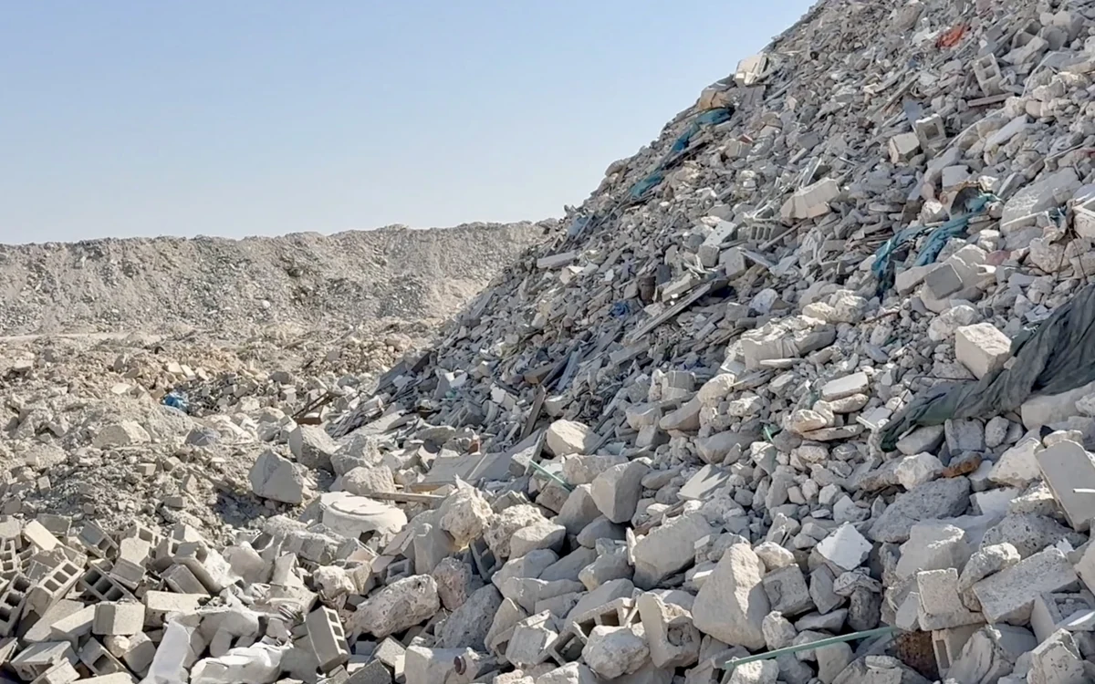 A mountain of demolition rubble under a clear sky, broken concrete, blockwork, slabs and torn netting tipped across the whole face of the tip and running away to the horizon