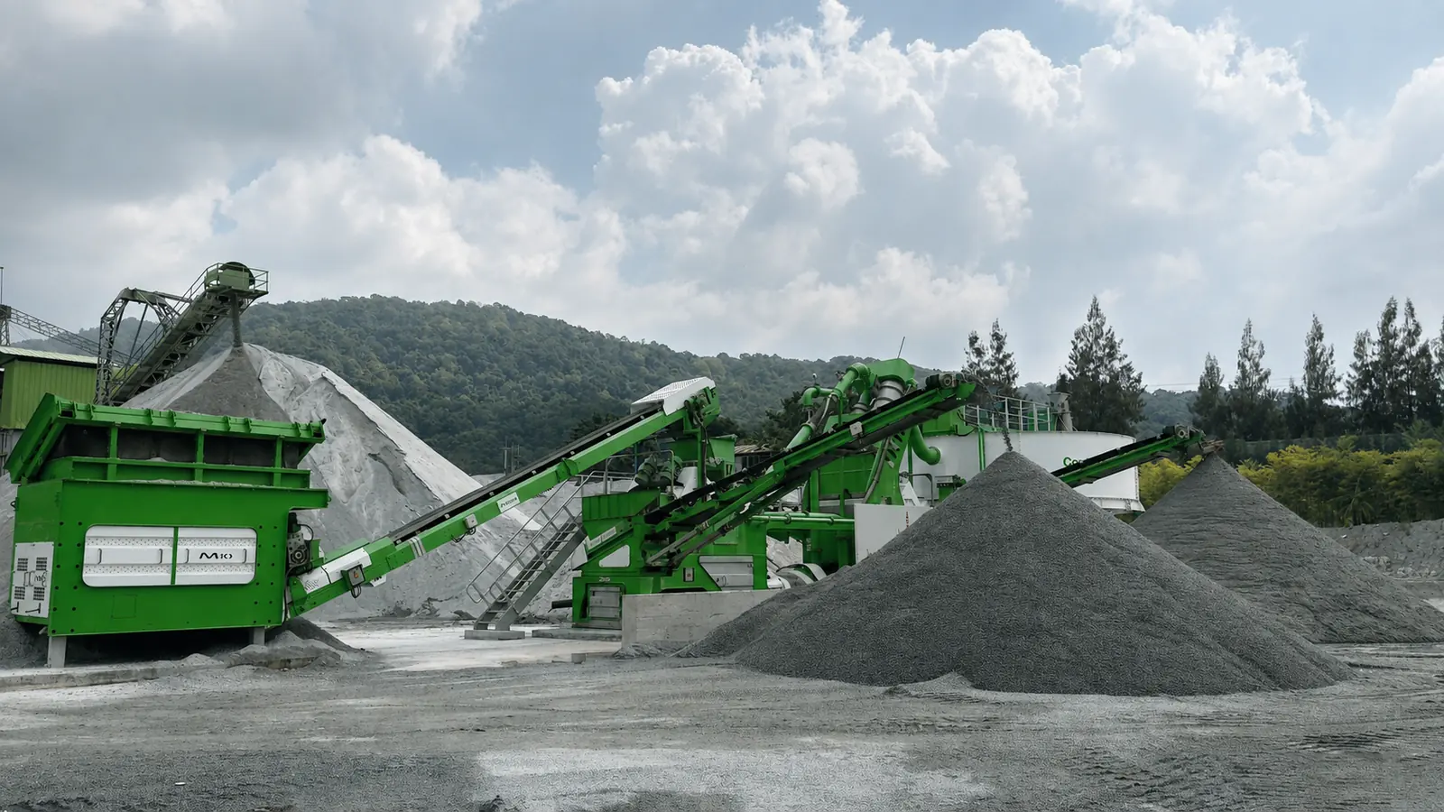 A CFlo Combo Alpha working in a quarry, feed hopper and conveyors in front of the thickener, with conical stockpiles of washed grey aggregate either side and forested hills behind