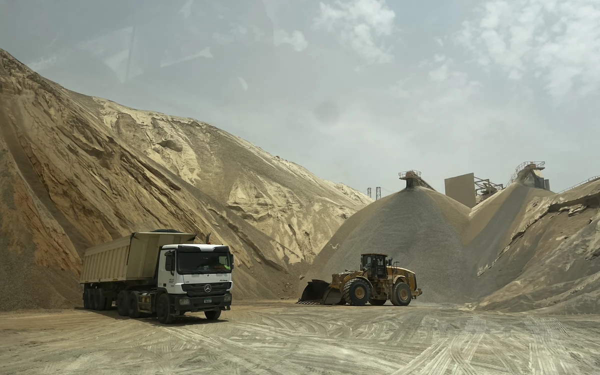 A quarry yard walled in by stockpiles, a long ridge of pale crusher dust on one side and conical piles of graded grey aggregate on the other, with a wheel loader and a tipper working between them and the crushing plant on the skyline