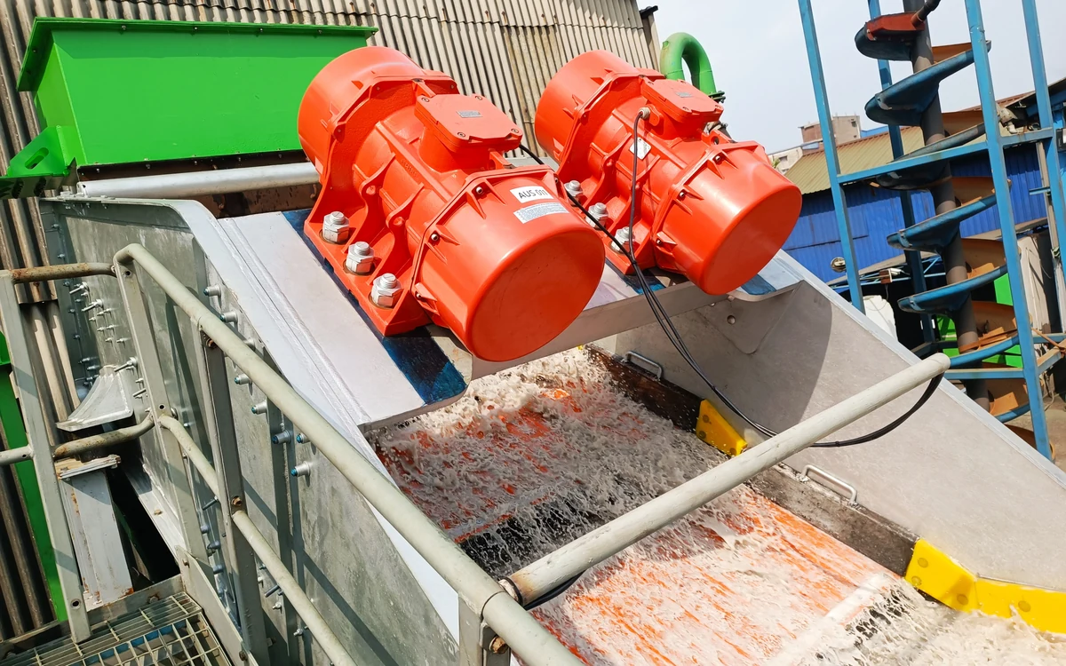 A pilot screen running under spray water, twin orange exciter motors bolted to the screen box and slurry washing down the deck below them, with a green feed chute at one side and a spiral classifier behind
