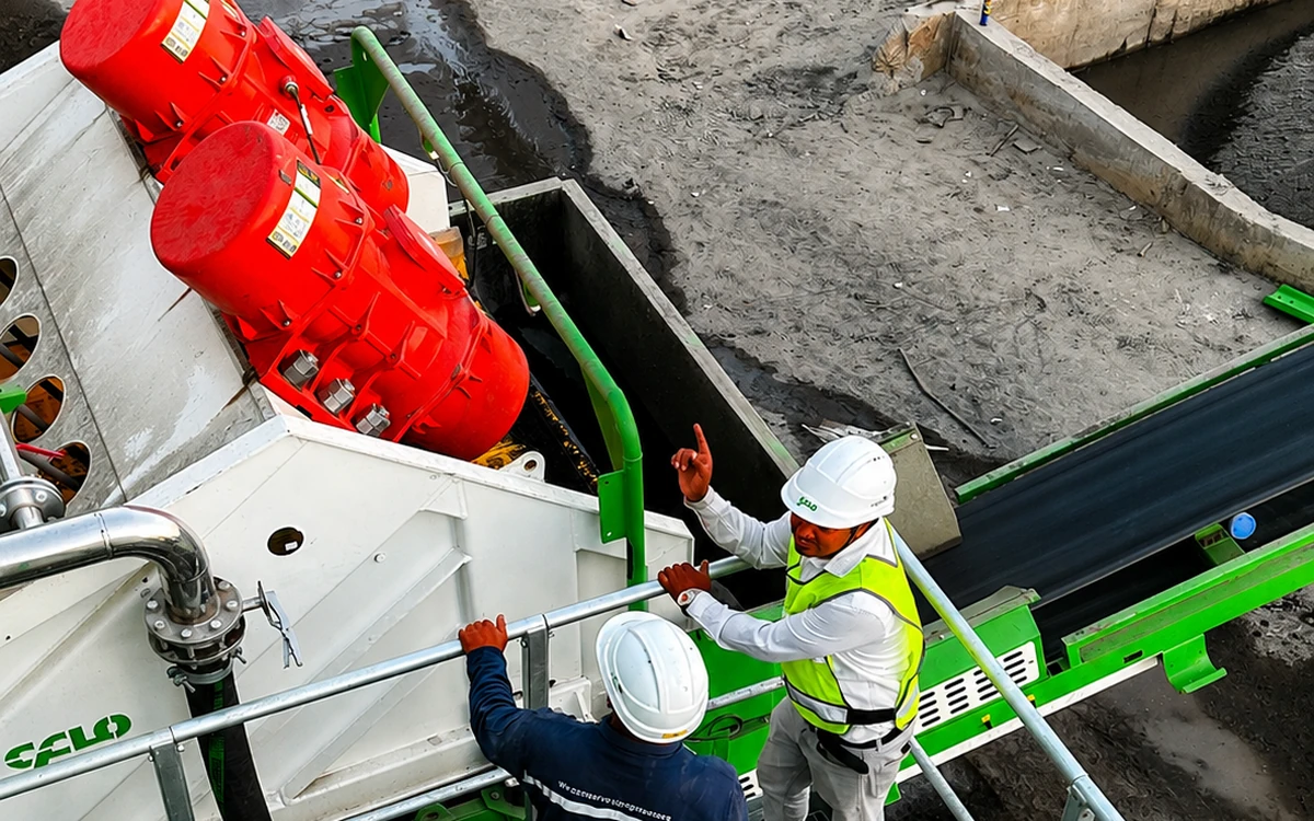 Two CFlo engineers in hard hats on the walkway of a Screenmax Dewatering & Sizing Screen during commissioning, one pointing along the deck, with the red twin exciters on the white screen box below them and the product conveyor running out across the site