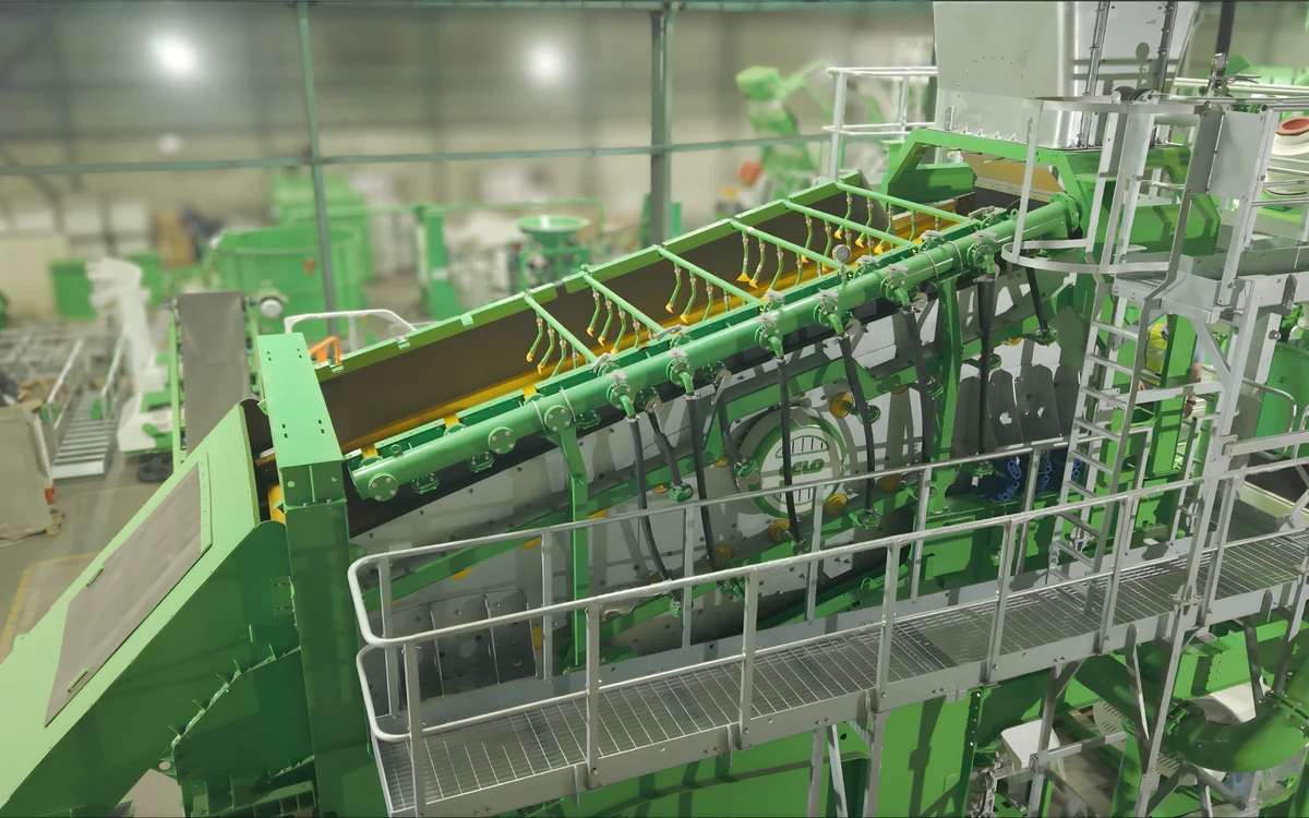 Spray bars along the top deck of a Screenmax G318 rinsing screen in the works, a row of yellow nozzles angled down onto the deck from the green manifold, with the screen box and its walkway below and the rest of the plant behind