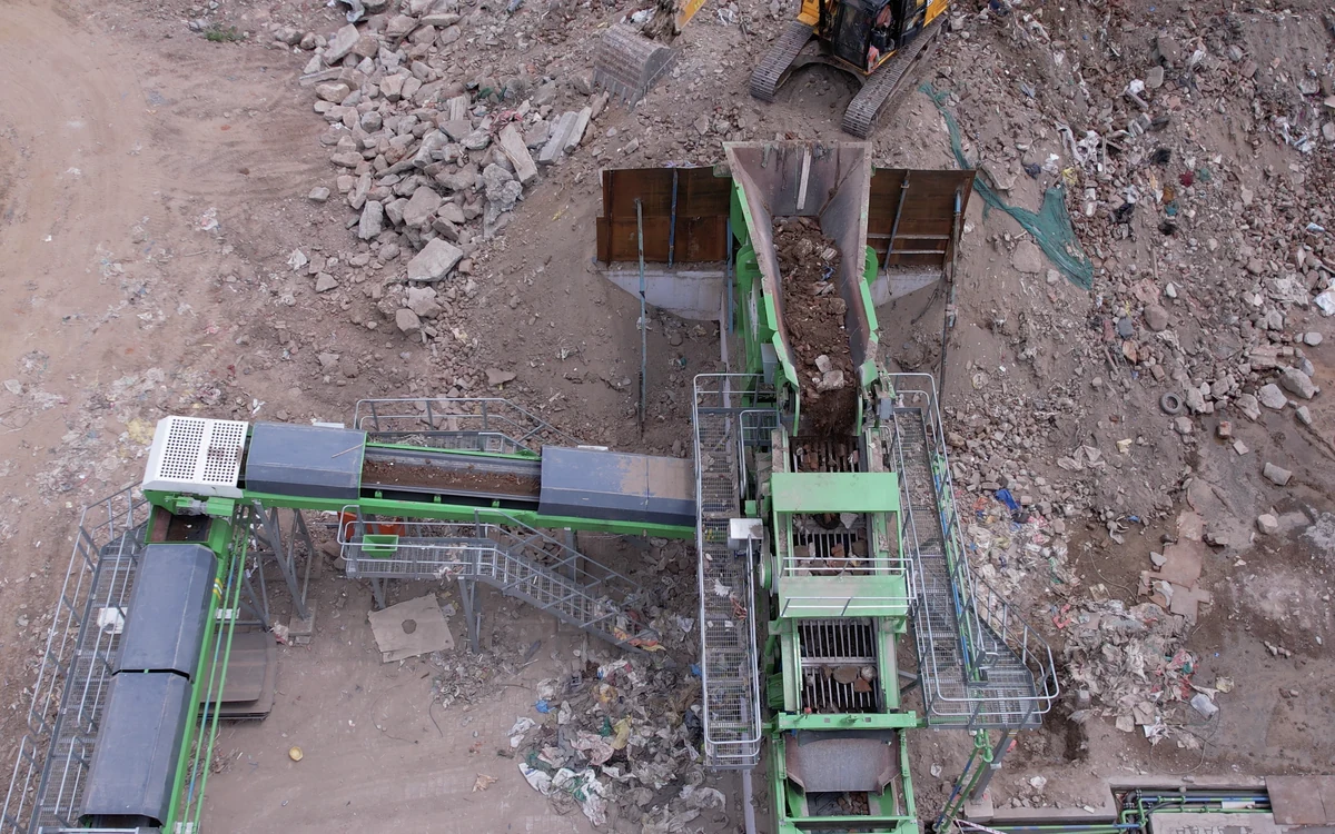 Overhead view of a Strato scalping screener on a demolition site, a tracked excavator tipping mixed rubble into the feed hopper, the grizzly deck below it and the under-pan and oversize conveyors running out to either side